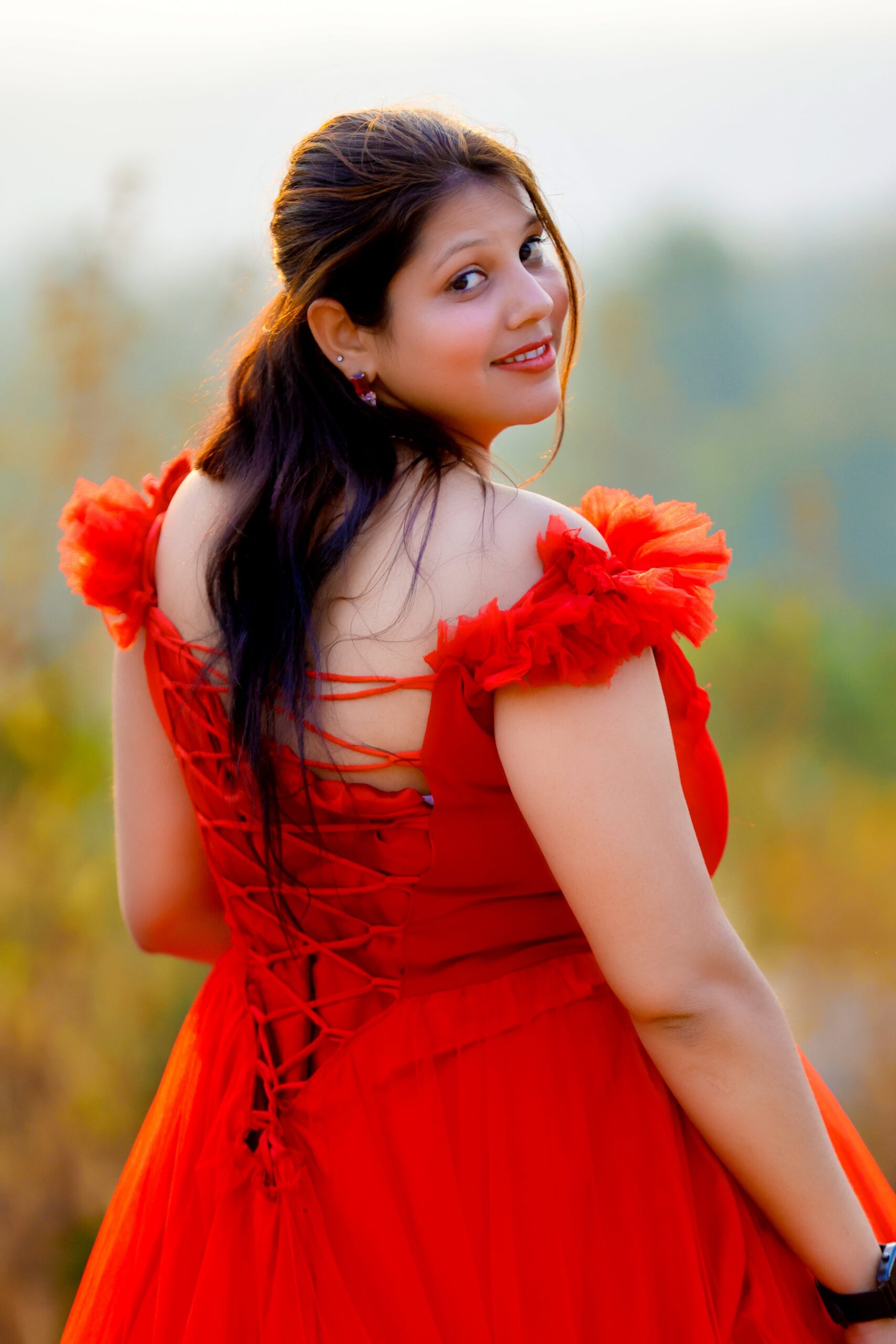 Woman in red off-shoulder dress with back lace-up in natural setting, looking over shoulder.